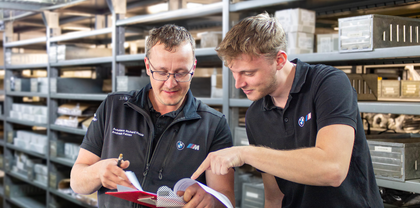 Azubi und Auszubildener zur Fachkraft für Lagerlogistik schauen gemeinsam in einem Lager auf Unterlagen auf einem roten Klemmbrett