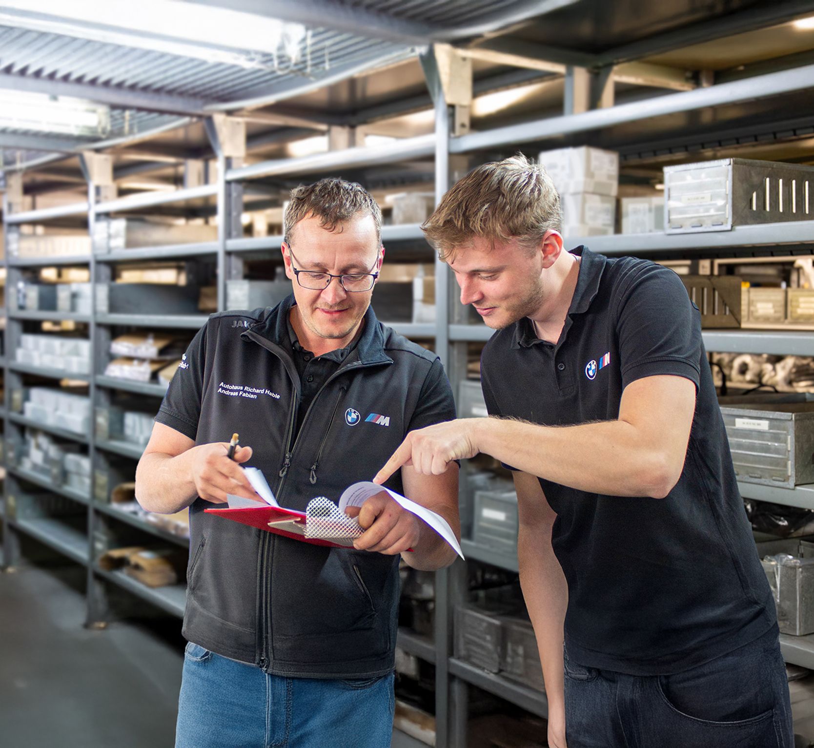 Azubi und Auszubildener zur Fachkraft für Lagerlogistik schauen gemeinsam in einem Lager auf Unterlagen auf einem roten Klemmbrett